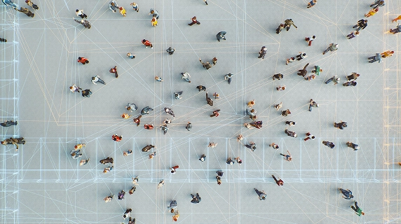 Top-down view of people walking in different directions on a pale surface, overlaid with a network of white and orange lines connecting various points, indicating movement tracking or spatial data analysis..