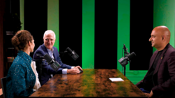 Three people engage in a podcast recording at a wooden table with professional microphones, against a green-lit background, creating a focused and lively atmosphere.