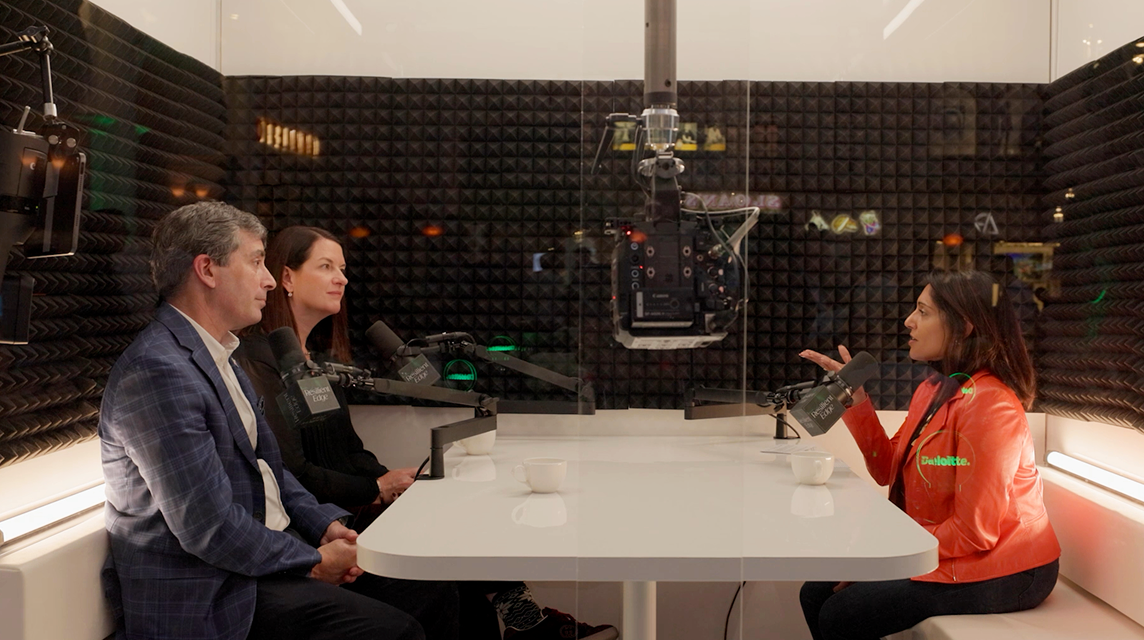 Three people engage in a podcast recording at a wooden table with professional microphones, against a green-lit background, creating a focused and lively atmosphere.