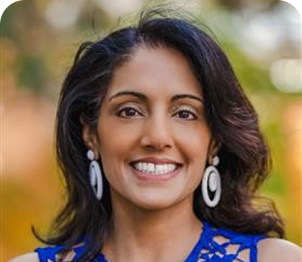 Person with shoulder-length dark hair wearing a bright blue sleeveless top, standing outdoors with soft greenery in the background.