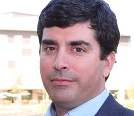 Person with short dark hair wearing a light blue collared shirt and a dark blazer, standing outside with a building in the background.