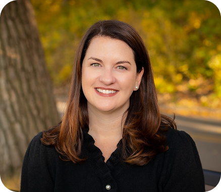 Person with long brown hair wearing a black top, seated outdoors on a bench with autumn foliage and a large tree trunk behind them.