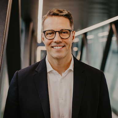 A person in a dark suit jacket and white dress shirt stands in a modern, well-lit corridor with glass walls and metal beams. 