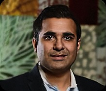 Person wearing a dark suit jacket over a light blue collared shirt, standing in front of a patterned background.