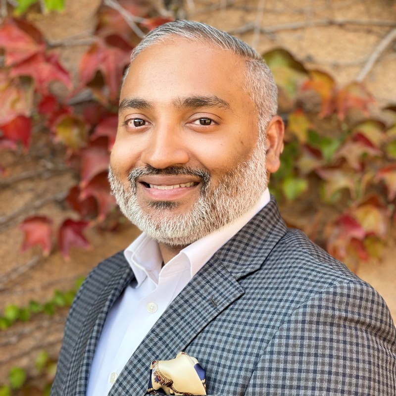 Person with short gray hair and beard, smiling, wearing a checkered blazer and white shirt with pocket square. Background of red and green ivy leaves.