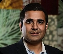 Person wearing a dark suit jacket over a light blue collared shirt, standing in front of a patterned background.