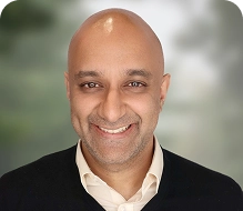 Person dressed in a white collared shirt under a dark sweater, standing against a soft-focus green and gray background.