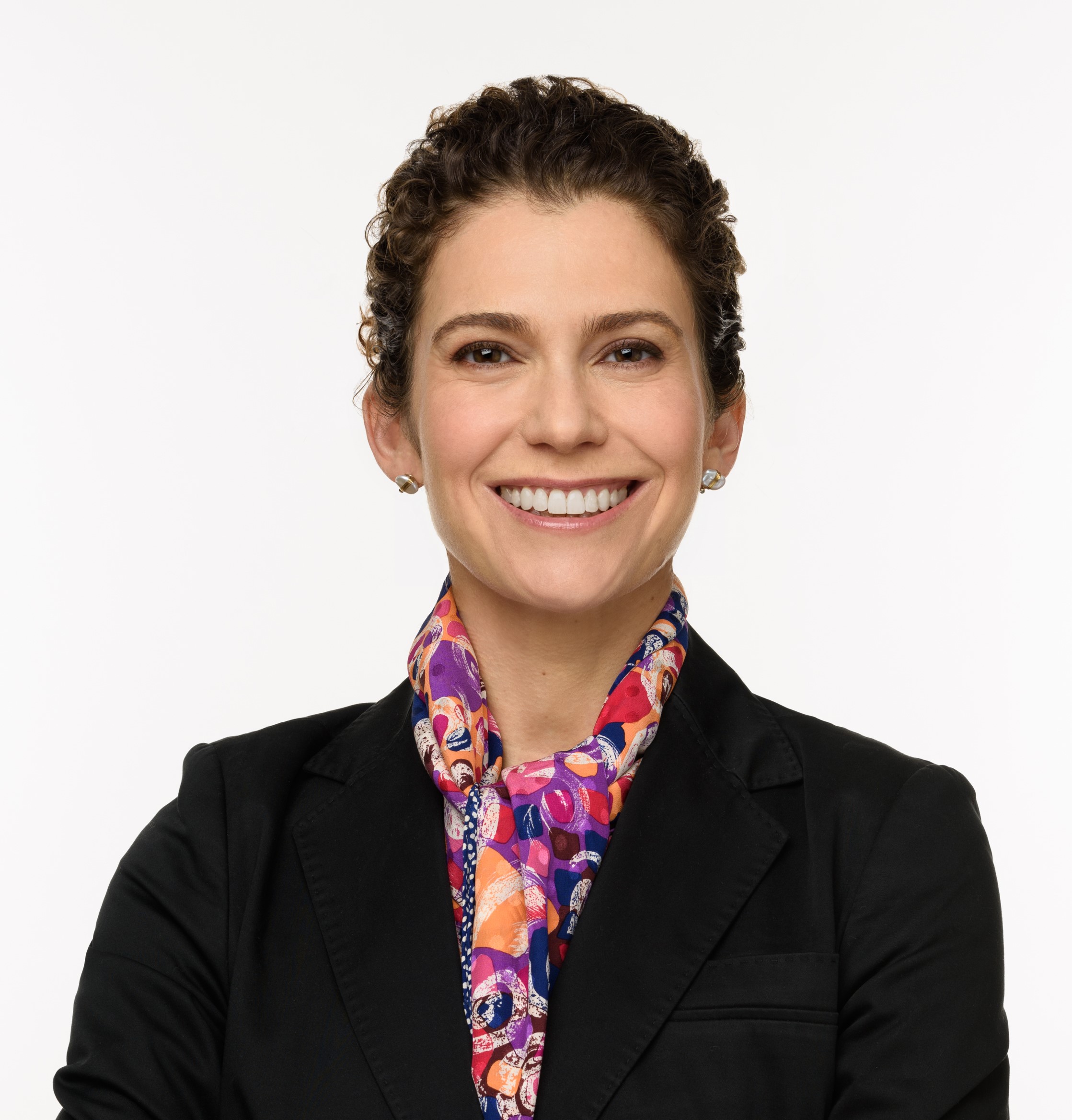 Person in a black suit jacket with a colorful patterned scarf, arms crossed, posed against a plain white background.